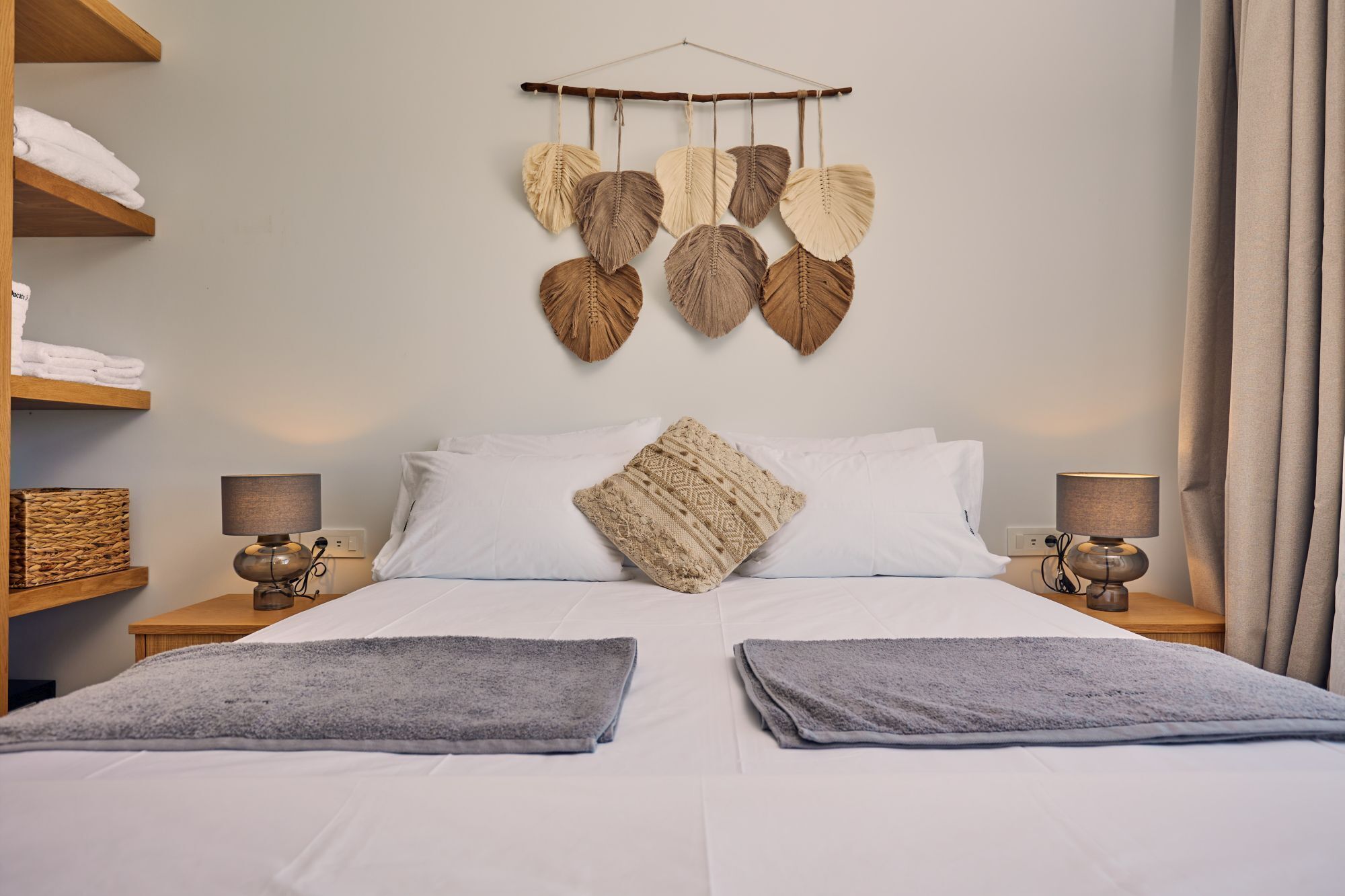 A neatly made bed with white linens, two gray towels, and a decorative wall hanging above.