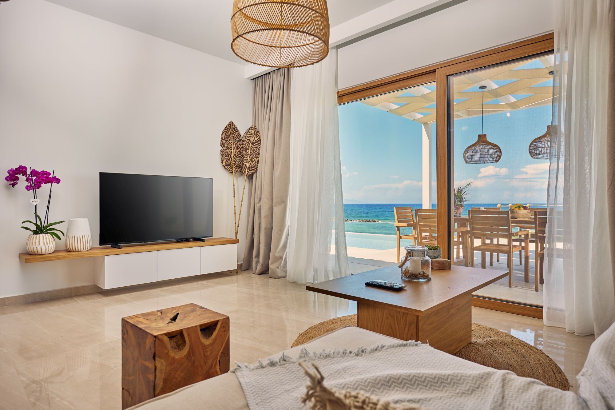A modern living room with a TV, wooden furniture, a view of the beach, and a dining area with outdoor seating.