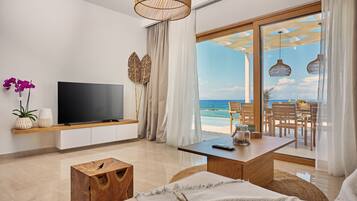 A modern living room with a TV, wooden furniture, a view of the beach, and a dining area with outdoor seating.