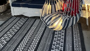 Ein Korbsessel, Sitzkissen mit Muster, ein hängender Makramee-Stuhl und ein Teppich mit Design auf einer Veranda mit Fenstern.