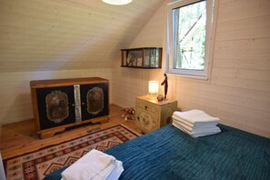Ein Schlafzimmer mit geneigter Decke, einem hölzernen Schminktisch, einem Nachttischchen mit Lampe, einem Hund und einem Fenster mit Blick auf Bäume.