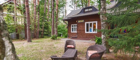 A wooden cabin with a porch, surrounded by tall trees and a grassy yard.