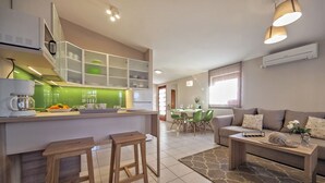 Eine moderne Küche mit einer zentralen Kochinsel, zwei Holzbarhocker, einem Essbereich mit grünen Stühlen und einem Wohnbereich mit Sofa und Couchtisch.
