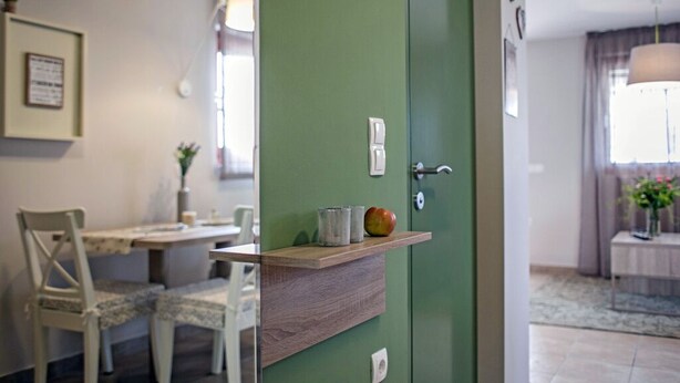 A green door with a small shelf above it, a white chair, and a table with a vase and a plant. - App. B - Grünes Appartment (Vir)