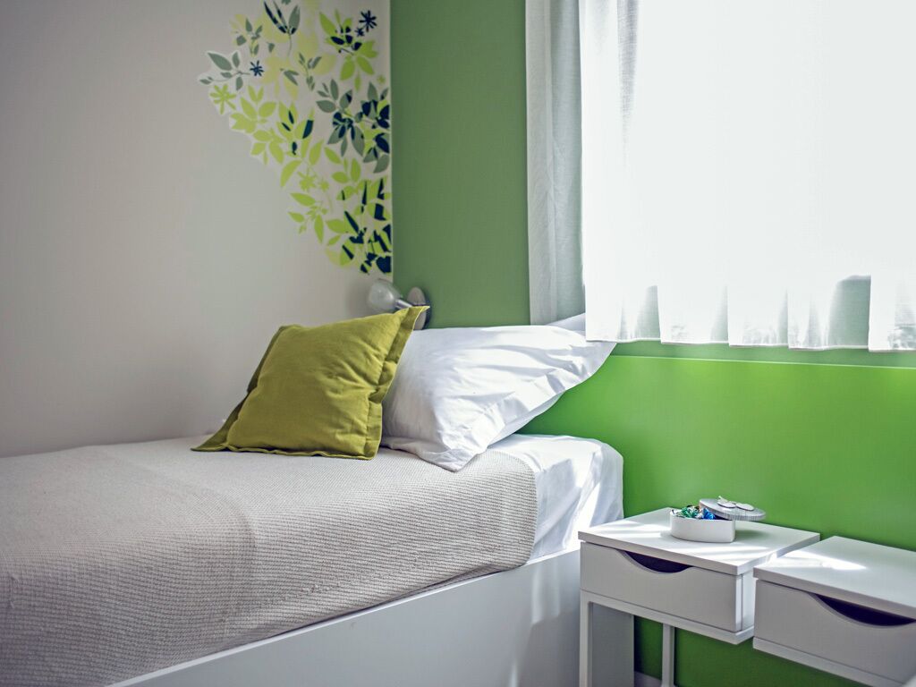 Un lit bien fait, aux couleurs blanches et vertes, une petite table de chevet avec une lampe et une fenêtre avec des rideaux.
