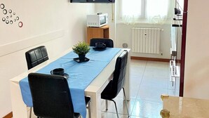 Uma sala de jantar com uma mesa comprida, cadeiras, uma TV na parede, um micro-ondas sobre uma mesinha e uma janela com cortinas.