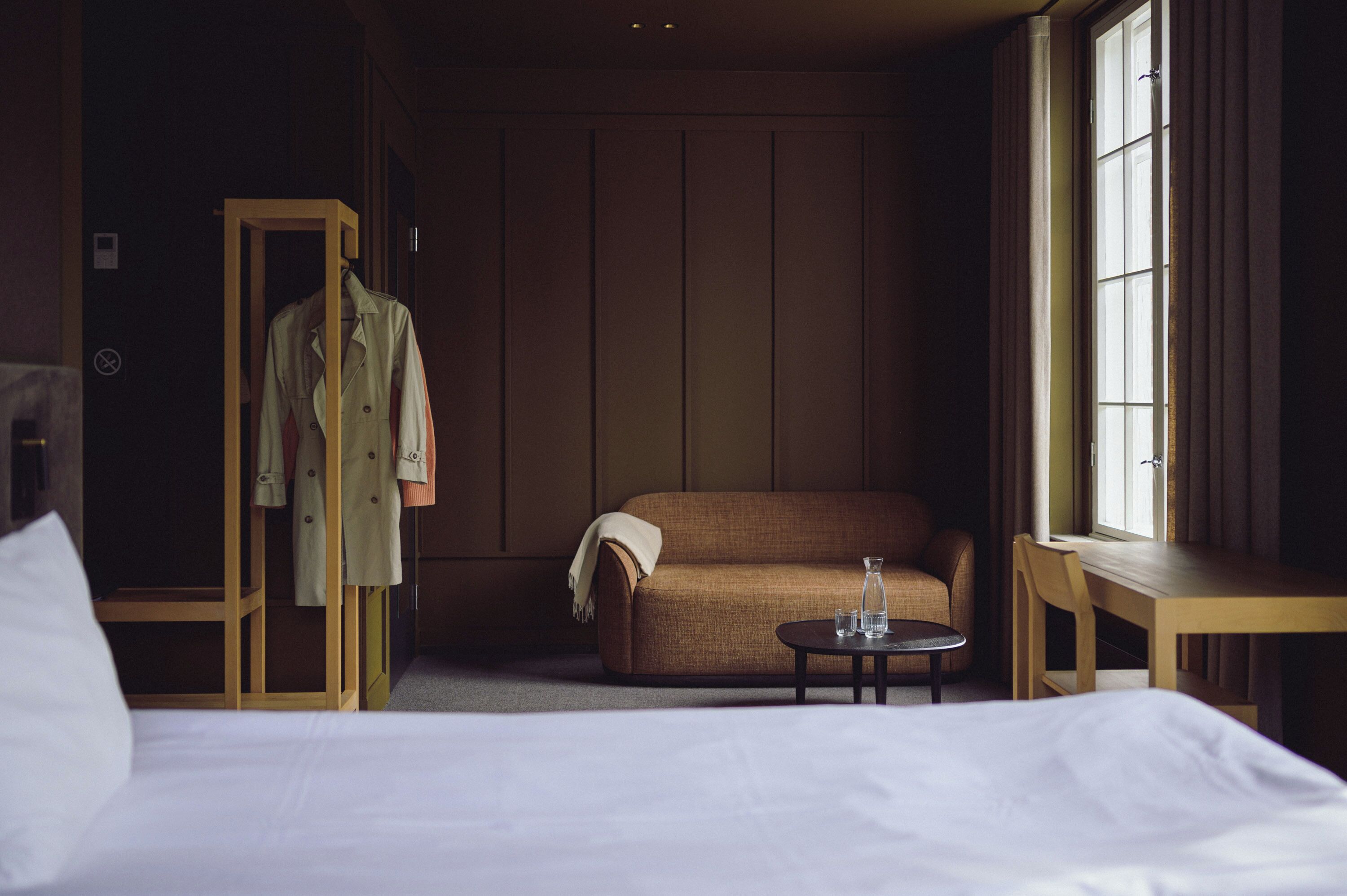 Habitación de hotel con cama, sofá marrón, armario de madera, mesita redonda con vasos y ventana con cortinas.
