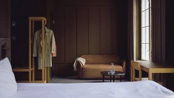 Habitación de hotel con cama, sofá marrón, armario de madera, mesita redonda con vasos y ventana con cortinas.