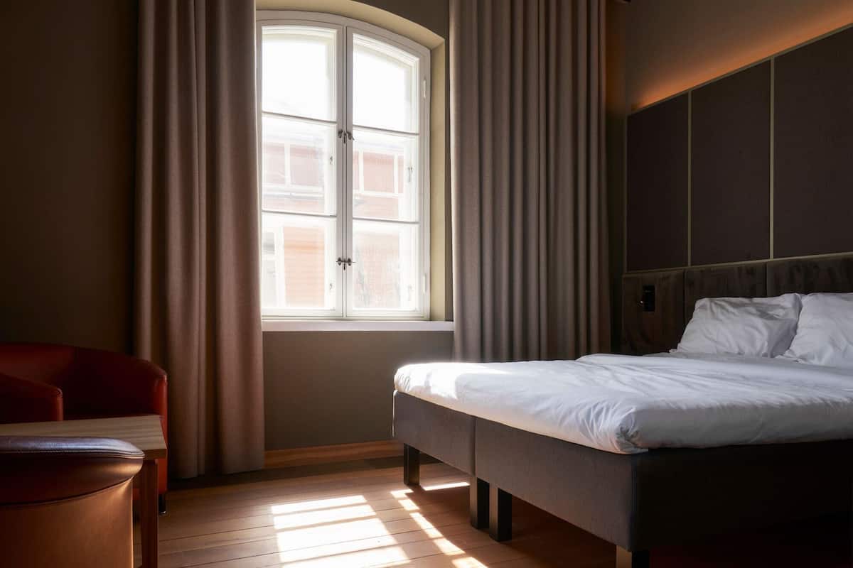 A hotel room with a bed, a window with curtains, a wooden floor, and a red chair.