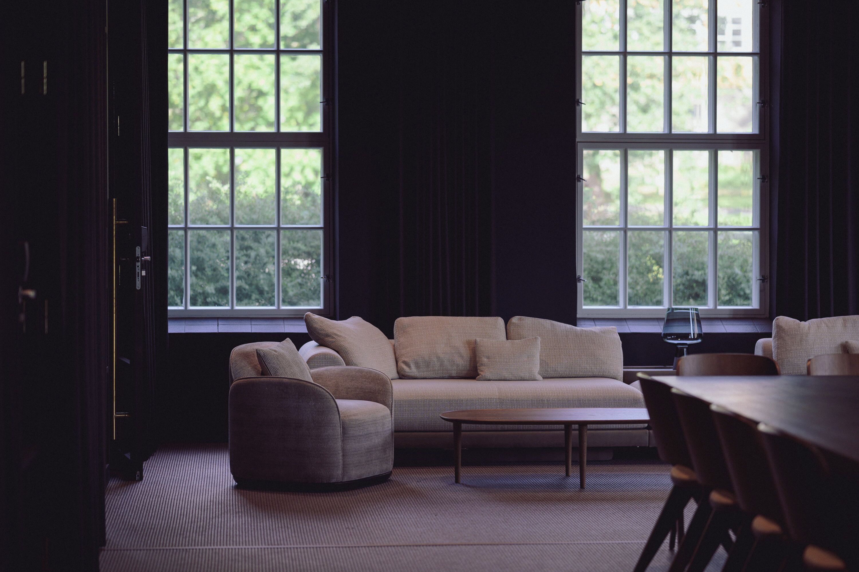 Una sala de estar moderna con sofá, sillón y mesa de centro. Amplias ventanas con vista a follaje exterior.