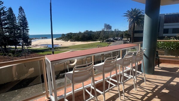 A balcony with white chairs, a red roof, and a view of a beach and ocean.