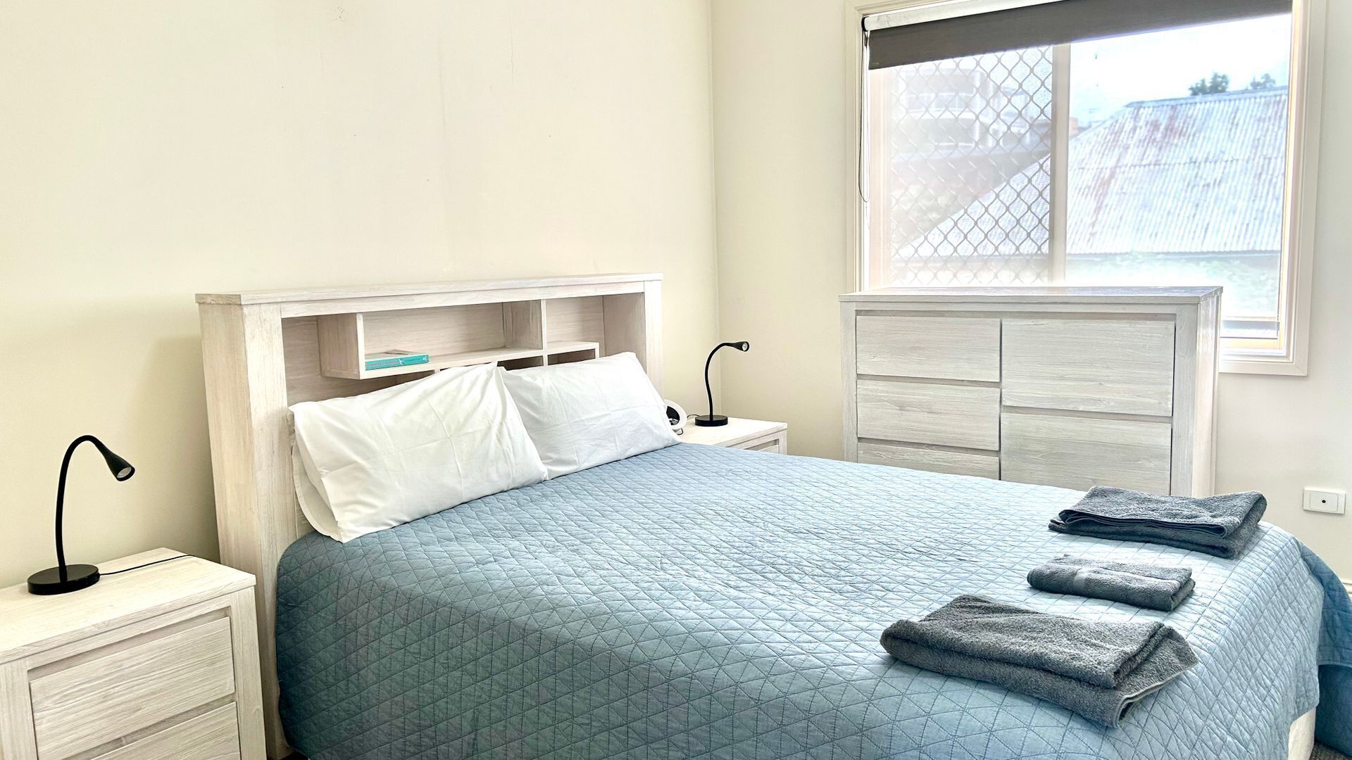 A neatly made bed with a blue quilt, a bedside lamp, a nightstand, and a window with a view of a fence.