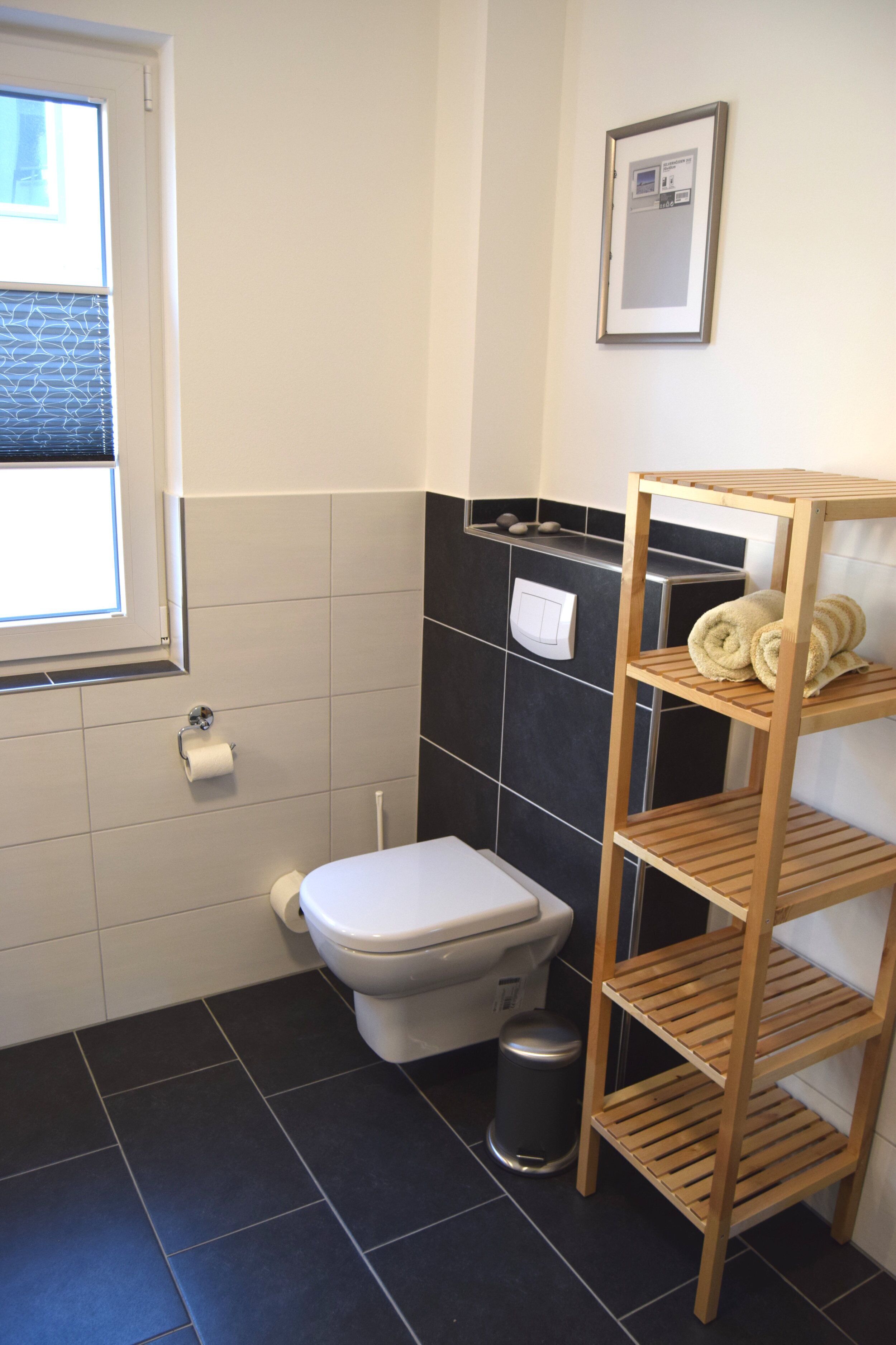 Un baño con inodoro, un estante con toallas, un cubo de basura y una ventana.