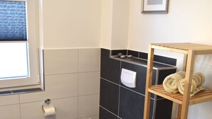 Un baño con inodoro, un estante con toallas, un cubo de basura y una ventana.