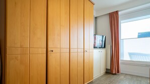 Una habitación con un armario de madera, un televisor sobre un mueble bajo y una ventana con cortinas.