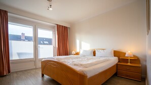 Un dormitorio con una cama de madera, dos mesitas de noche con lámparas, una puerta corredera de vidrio y vistas a edificios.
