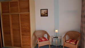 Une pièce comprenant une armoire en bois, deux fauteuils en osier avec des coussins à carreaux, une petite table avec une lampe et un tableau encadré accroché au mur.