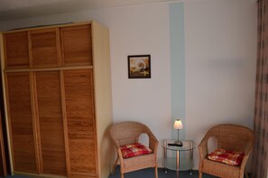 A room with a wooden wardrobe, two wicker chairs with plaid cushions, a small table with a lamp, and a framed picture on the wall.