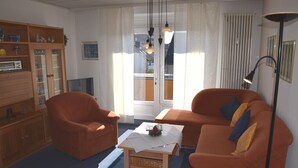 Un salon comprenant un canapé, un fauteuil et une table basse, un meuble en bois et une grande fenêtre avec des rideaux.