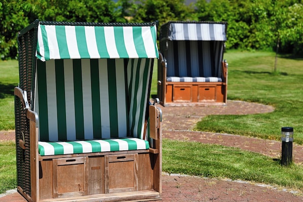 Deux fauteuils de jardin en bois, dotés de coussins à rayures, l’un vert et blanc, l’autre bleu et blanc, sont placés sur un chemin pavé de briques, dans une zone herbeuse.