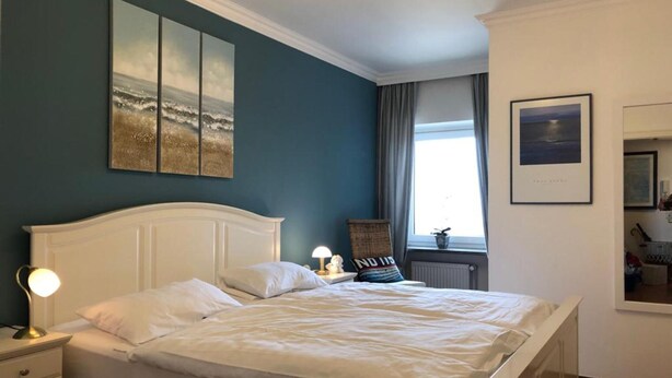 A bedroom with a bed, bedside lamps, a window with curtains, and artwork on the walls. - Büsumer Kuschelnest mit Meerblick (Büsum)