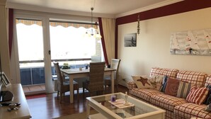 A living room with a sofa, a glass coffee table, a dining area with wicker chairs, and a view through a sliding glass door. - Büsumer Kuschelnest mit Meerblick (Büsum)