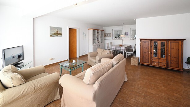 Ein Wohnzimmer mit beigefarbenen Sofas, einem Glastisch, einem Holzschrank und einem Essbereich mit Tisch und Stühlen.