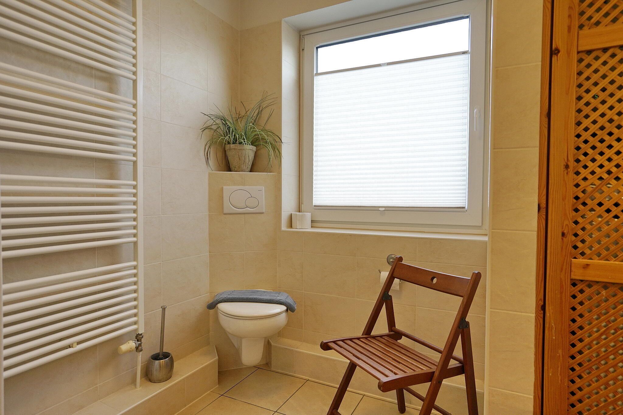 Un baño pequeño con inodoro, radiador, ventana y una silla de madera.