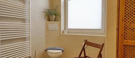 Un baño pequeño con inodoro, radiador, ventana y una silla de madera.