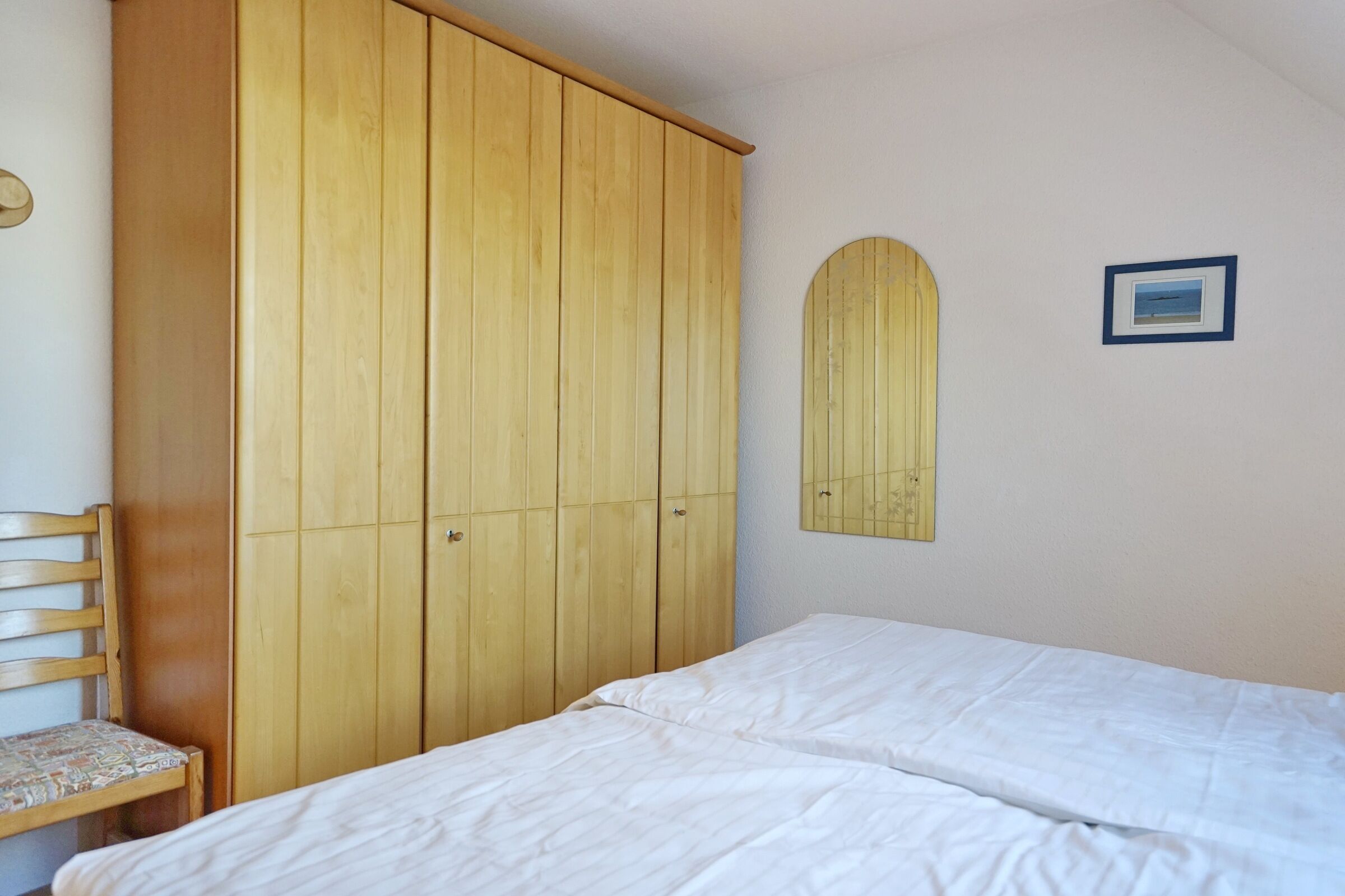 A bedroom with a wooden wardrobe, a bed, a chair, and a wall-mounted mirror.