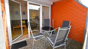 Et lille udendørs opholdsområde med et bord og to stole, en skydedør der fører til et indvendigt rum, og en orange væg med struktur.