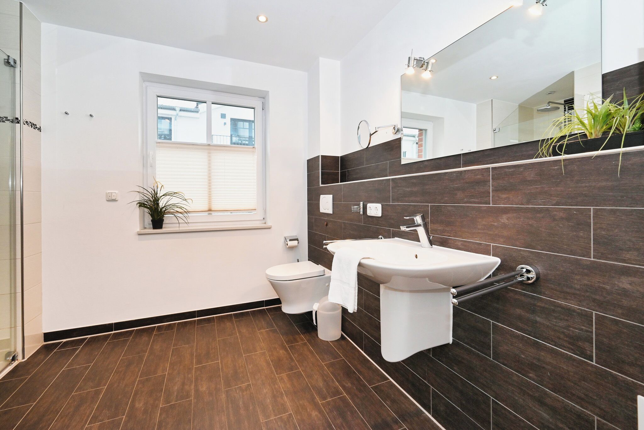 Een moderne badkamer met een wit toilet, een wastafel en een grote spiegel. Er is een raam met zonwering en een plant in een pot op het raamkozijn.