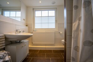 Une salle de bain moderne avec un lavabo blanc, un miroir et des toilettes. On y trouve une fenêtre avec des stores, un rideau de douche et un radiateur.