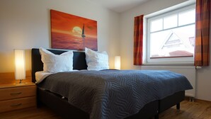 A bedroom with a bed, bedside lamps, a painting of a sunset, a window with curtains, and a radiator. - Haus Meereskönig Wohnung 2 (Büsum)