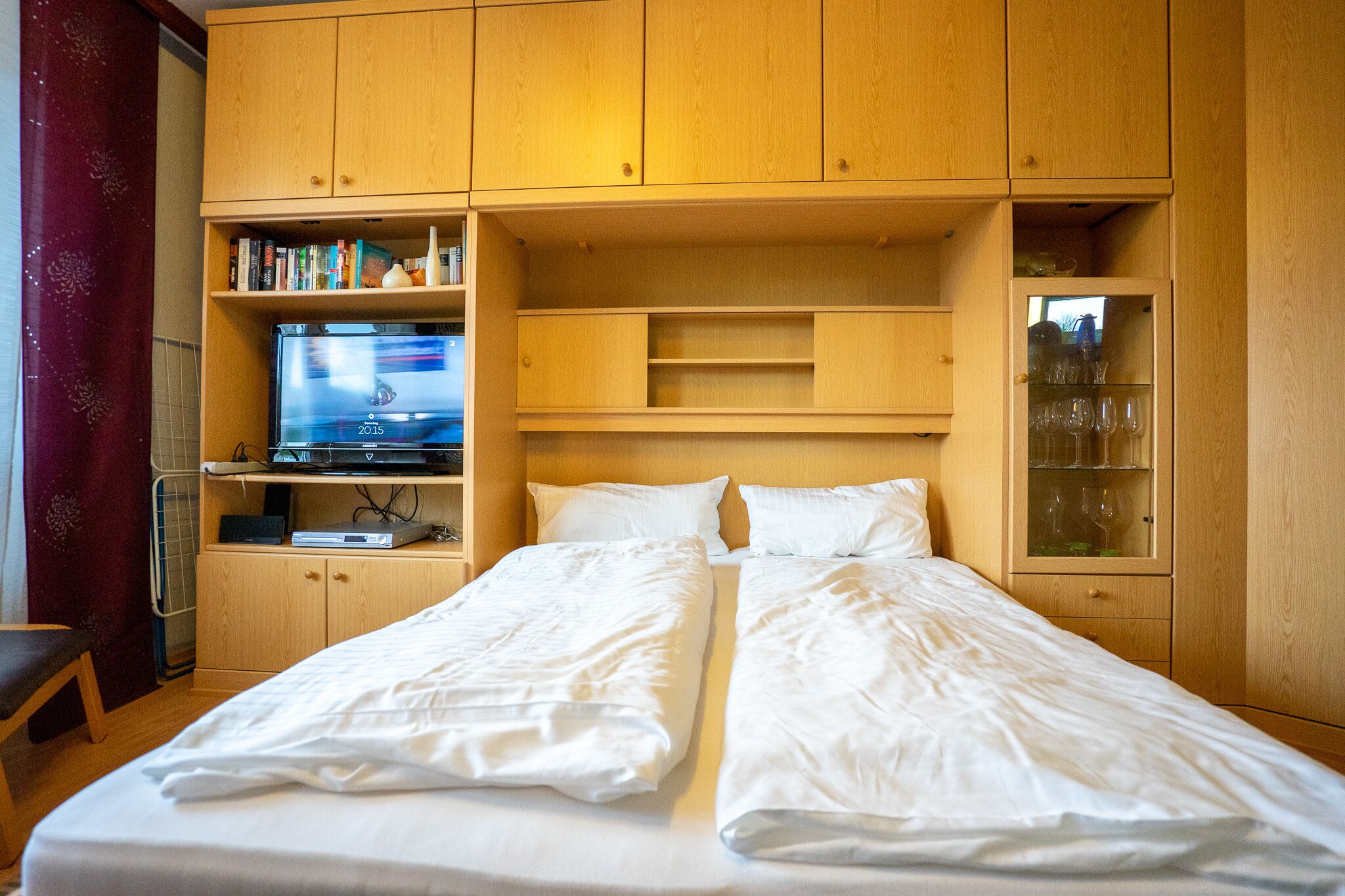 A bedroom with a bed, wooden cabinets, a television, and a shelf with books and glasses.