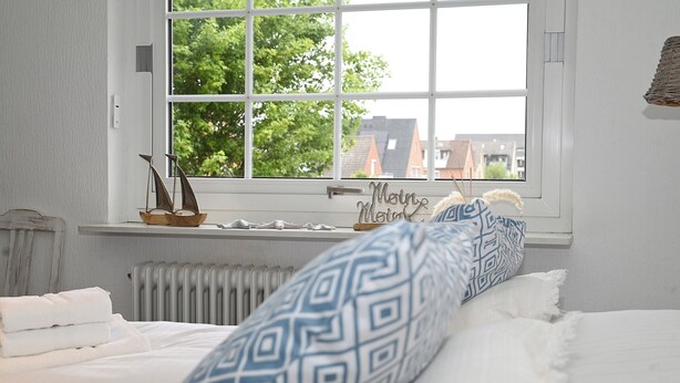 Ein Schlafzimmer mit einem Bett, einem Fenster mit Blick auf Gebäude, einem Heizkörper und einer kleinen Ablage mit Dekoration.