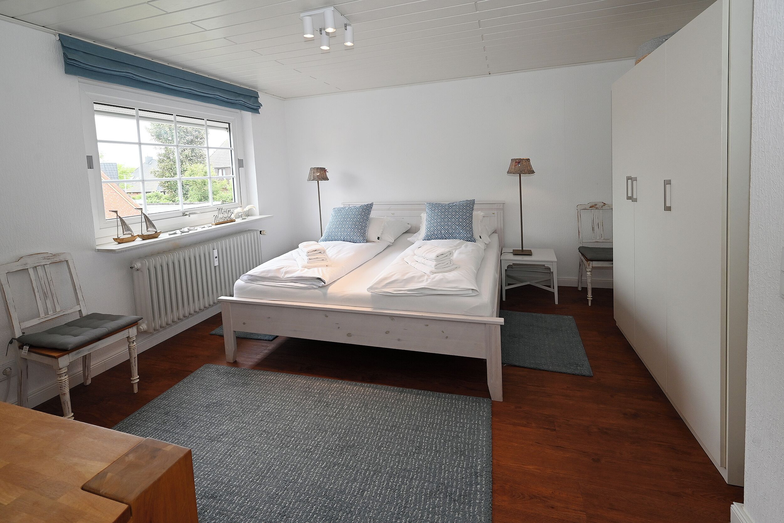 Quarto com cama, mesas de cabeceira, janela com cortinas, radiador e um banco de madeira.