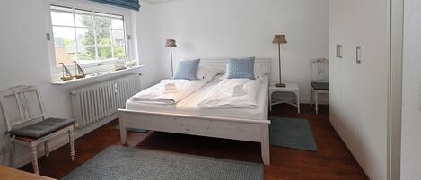 Quarto com cama, mesas de cabeceira, janela com cortinas, radiador e um banco de madeira.