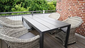 Eine Terrasse mit Tisch und Korbstühlen, eine Ziegelwand und ein Hecken im Hintergrund.