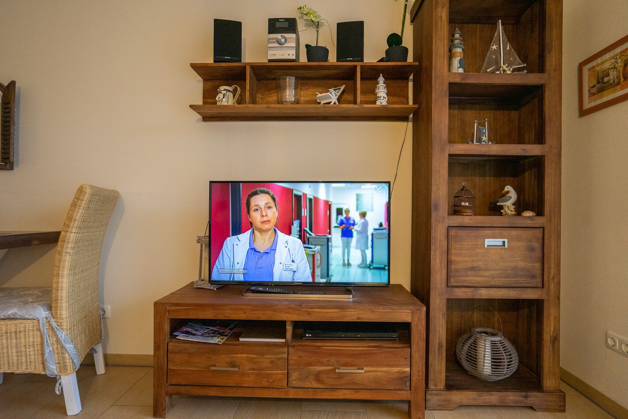 Un salon avec un téléviseur diffusant l’image d’une personne portant une blouse de laboratoire, un fauteuil en osier, un meuble de rangement en bois avec des étagères et un panier en osier.
