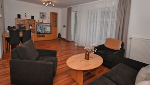 A modern living room with a wooden floor, two dark sofas, a round wooden coffee table with a vase, and a dining area with a wooden table and chairs. - Haus Schäfer Wohnung 1 in Ruhiger Lage (Büsum)