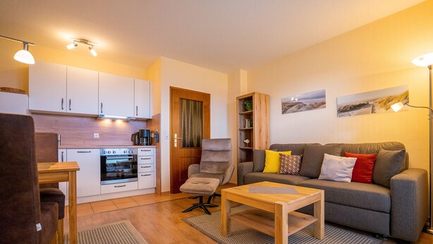 Een gezellig, compact verblijf met een kitchenette, een grijze bank met kleurrijke kussens, een houten bijzettafeltje en een fauteuil.