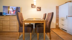 Een eethoek met een houten tafel en vier stoelen, een televisie waarop iemand te zien is, en twee ingelijste foto's aan de muur.