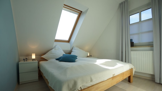 Een slaapkamer met een schuine plafond, een bed met witte beddengoed, twee nachtkastjes met lampen, een raam met gordijnen, en een radiator.