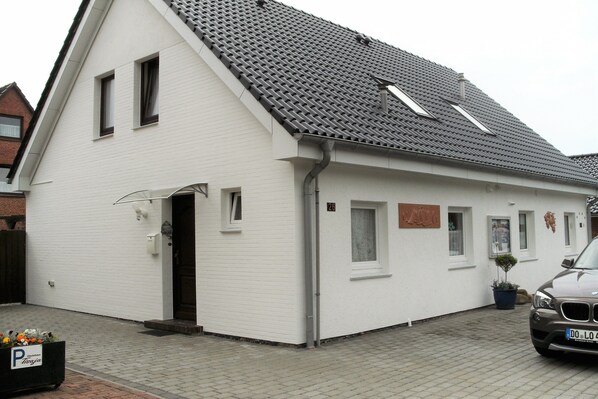 A white building with a grey tiled roof, a visible door, and a parked car. - Haus "reinhard" in Ruhiger Lage Direkt am Wäldchen (Büsum)