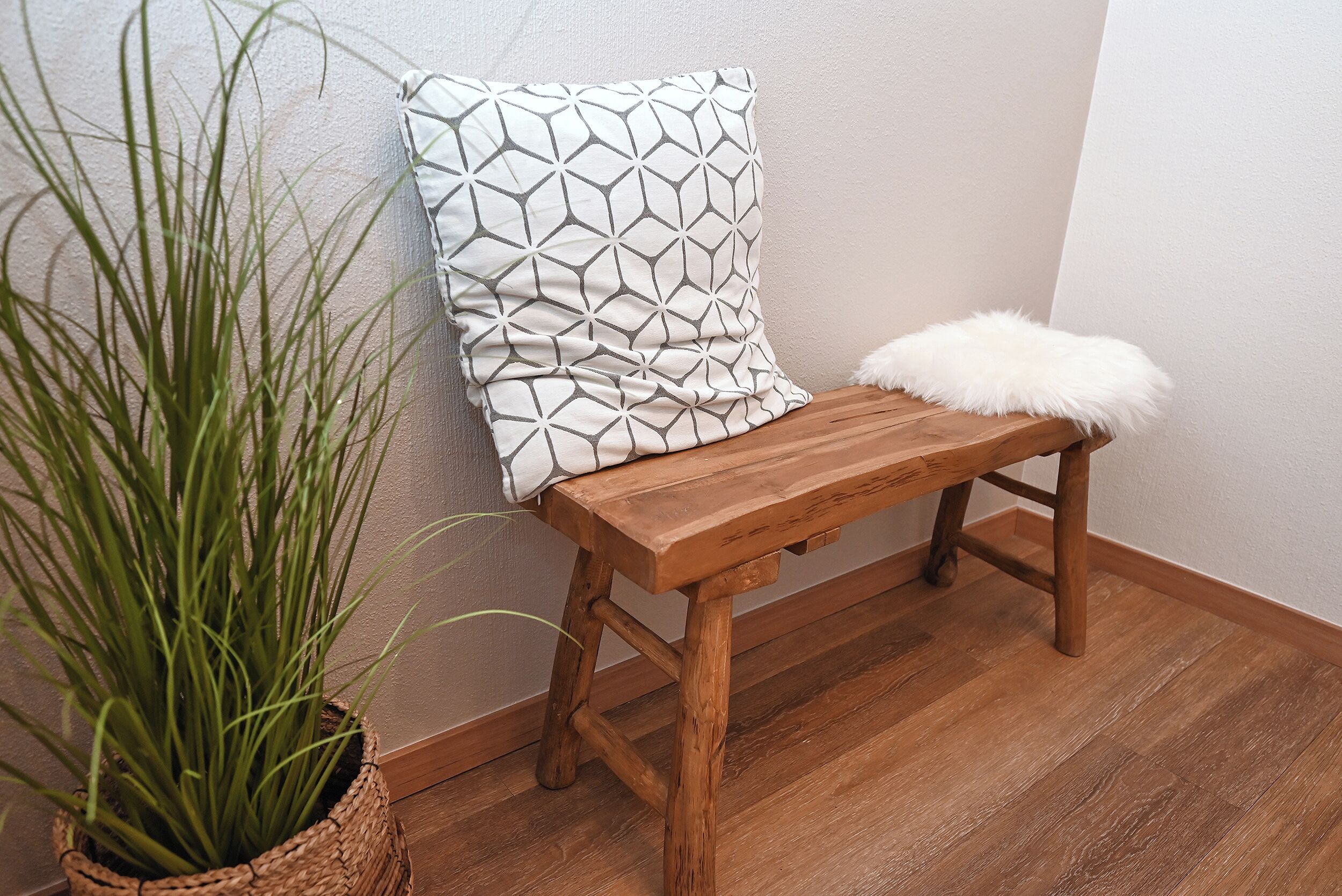Eine Holzbank mit einem Kissen in geometrischem Muster und einem flauschigen weißen Sitzkissen, neben einem Blumentopf mit langen, grünen Blättern.