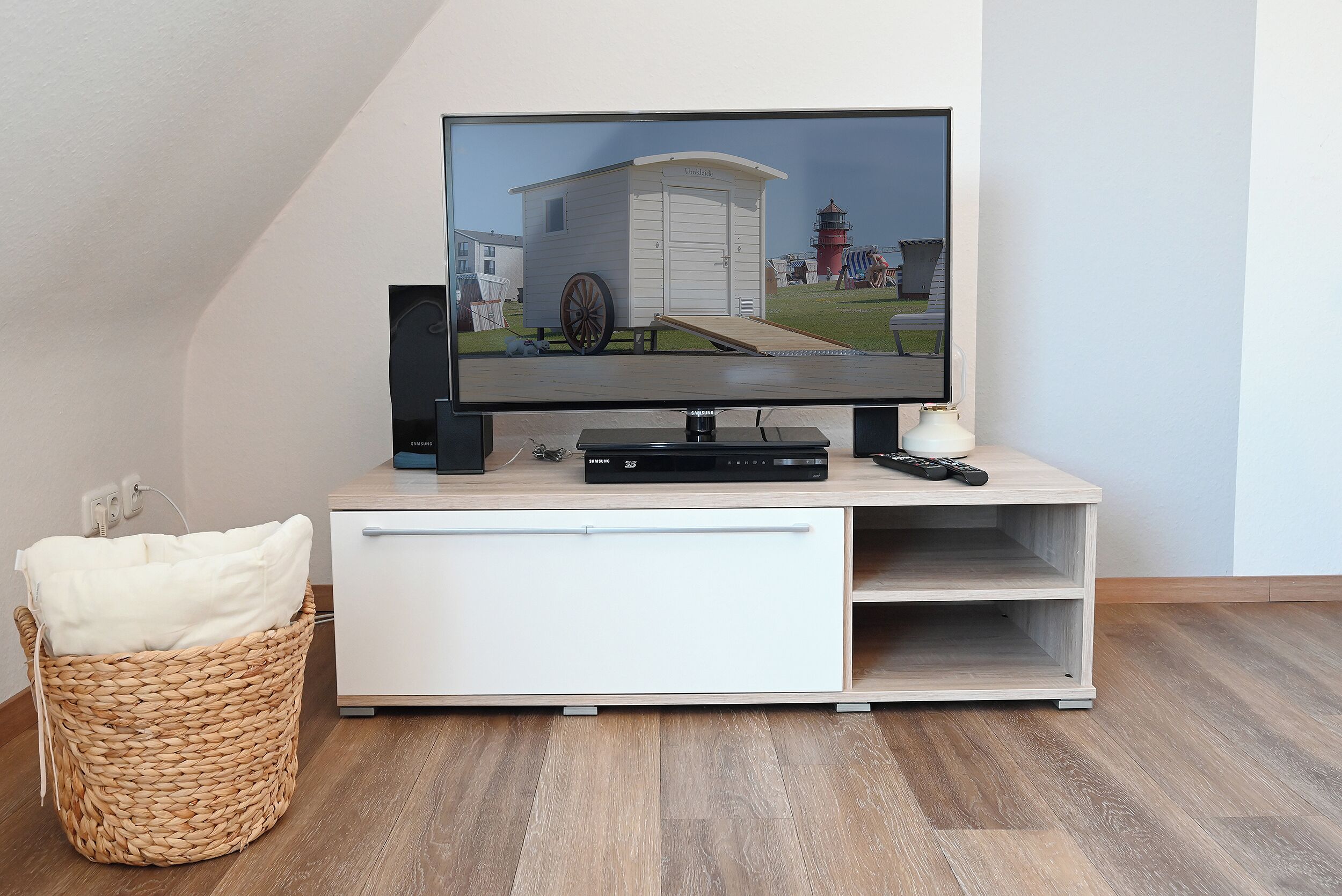A television on a wooden stand with a remote, a white cabinet below, and a wicker basket with a pillow.