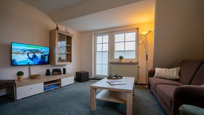 A living room with a television, a wooden coffee table, a brown sofa, a floor lamp, and a window with blinds.