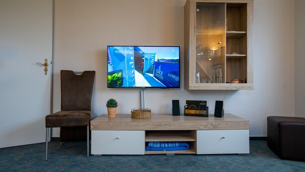 A modern living room with a flat-screen TV mounted on the wall, a wooden cabinet with glass doors, a brown upholstered chair, a small potted plant, and a white entertainment unit with drawers.
