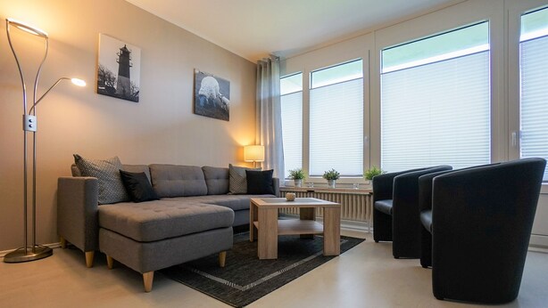 A modern living room with a grey sectional sofa, a small wooden coffee table, and black armchairs. There are two floor lamps, a radiator, and large windows with blinds. - Wohnung Eil 809 im Herzen von Büsum (Büsum)